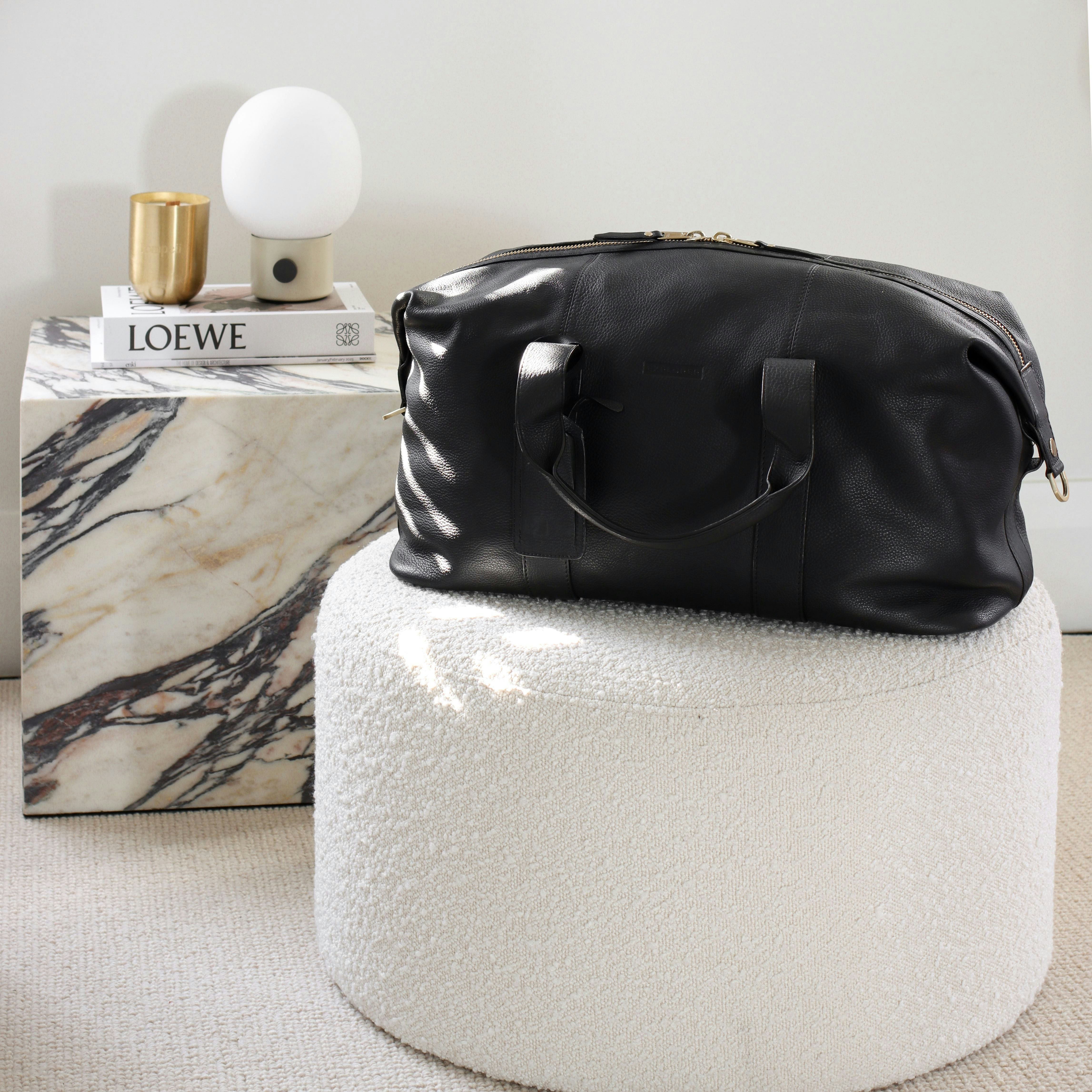 Explorer Weekender Leather Duffle in black on top of a white footstool in a room.