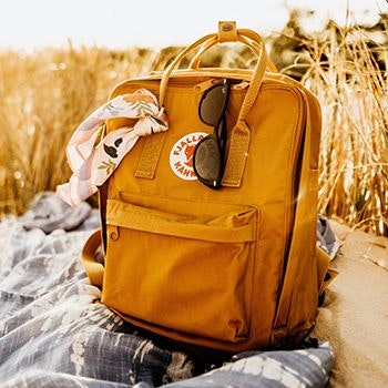 Fjallraven Kanken Backpack in Ochre on the beach, with sunglasses and scarf tied to it