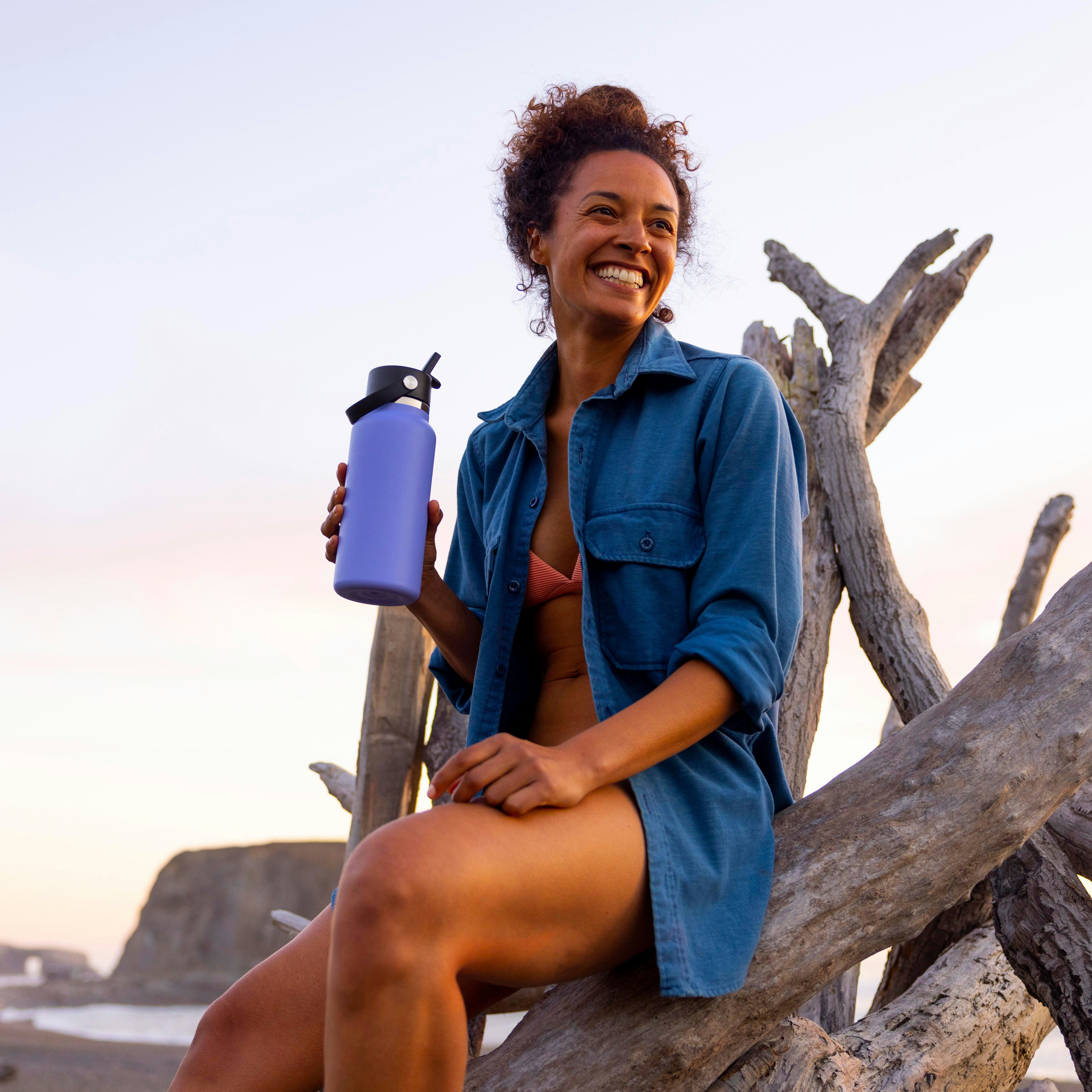 Women smiling with her purple Hydro Flask drink bottle at the beach at sunset sitting on a log. 